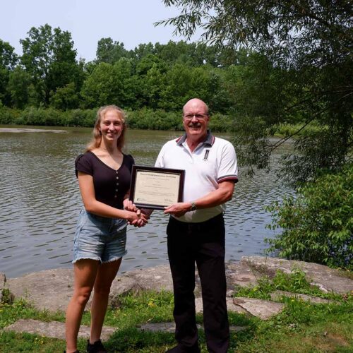 A young woman and man stand in front of water holding a certificate