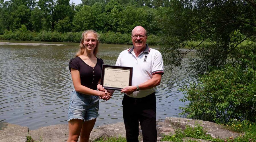 A young woman and man stand in front of water holding a certificate