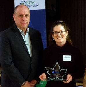 A man and a woman standing together. The woman is holding a leaf shaped award.